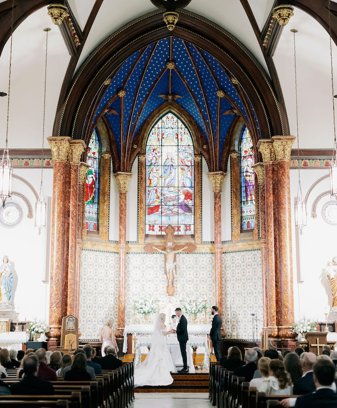 A bride and groom stand at the altar exchanging vows inside a church with stained glass windows. 