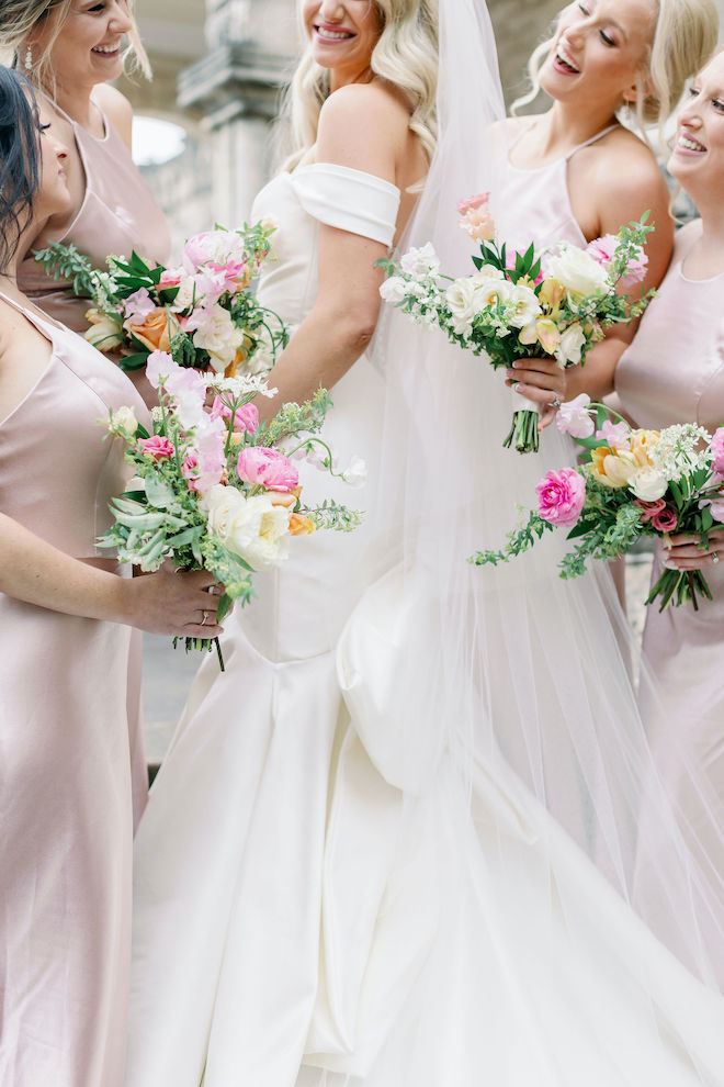 A bride wearing a white wedding dress and her bridesmaids wearing blush pink gowns hold their pastel wedding bouquets as the laugh. 
