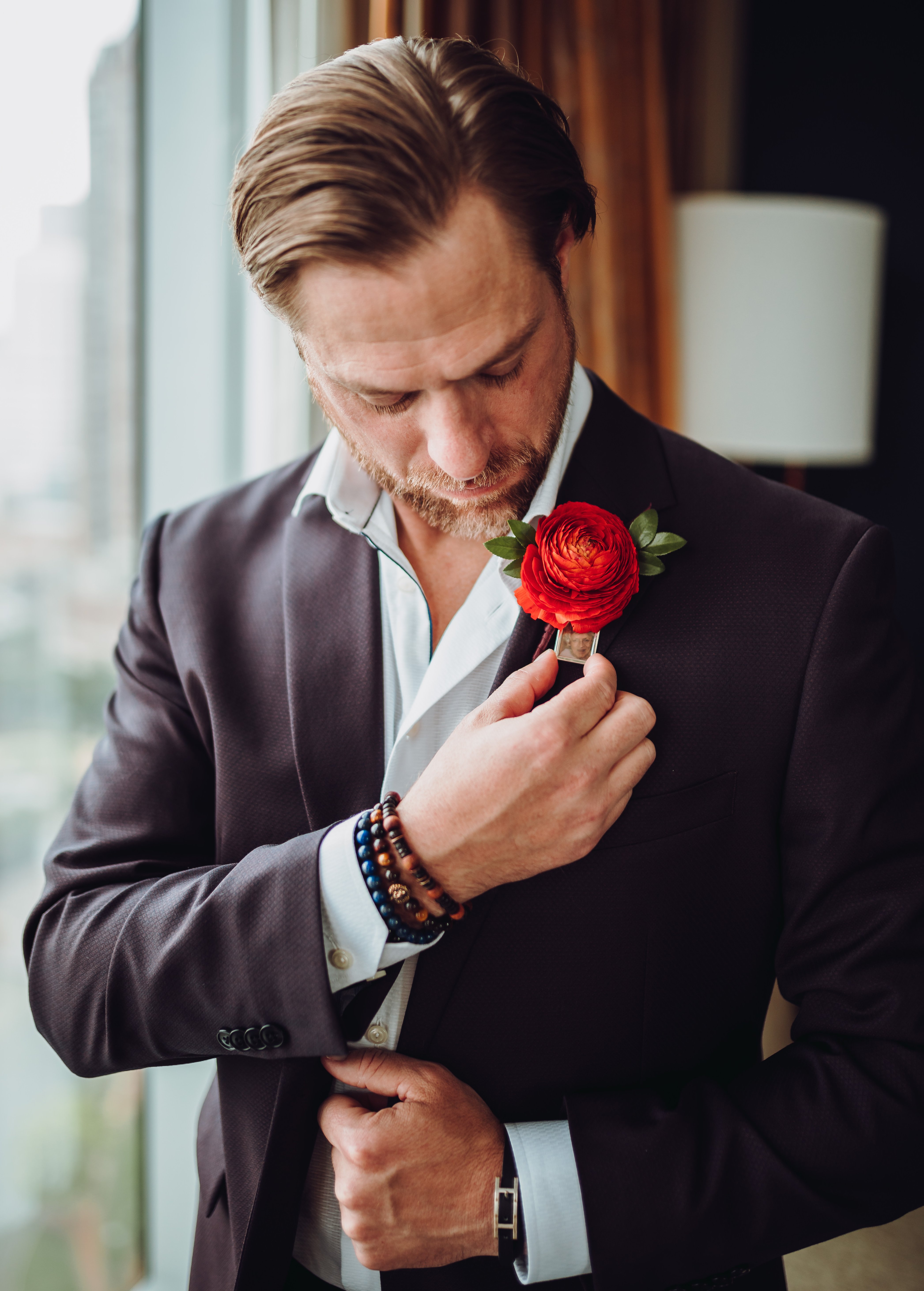 A groom looks down at his boutonierre which has a bright red Ranunculus and a photo of his late mother.
