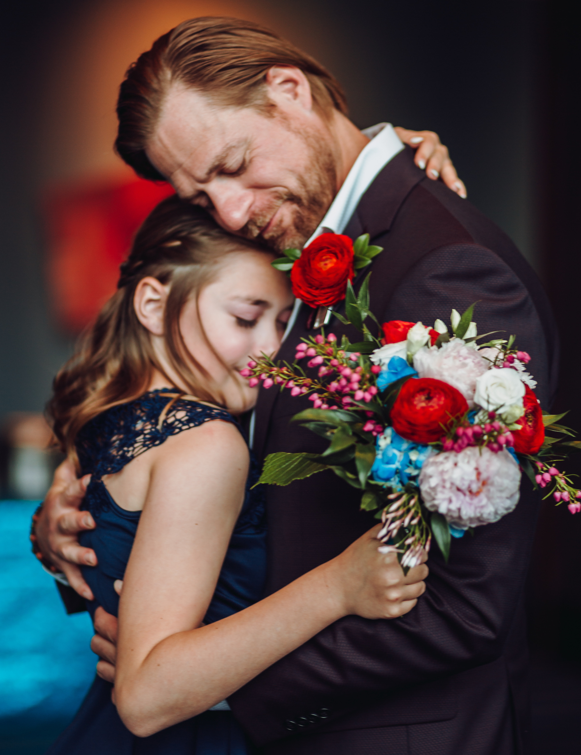 A groom hugs his daughter before his wedding ceremony. She is holding a colorful bouquet full of red, blue and peach flowers.