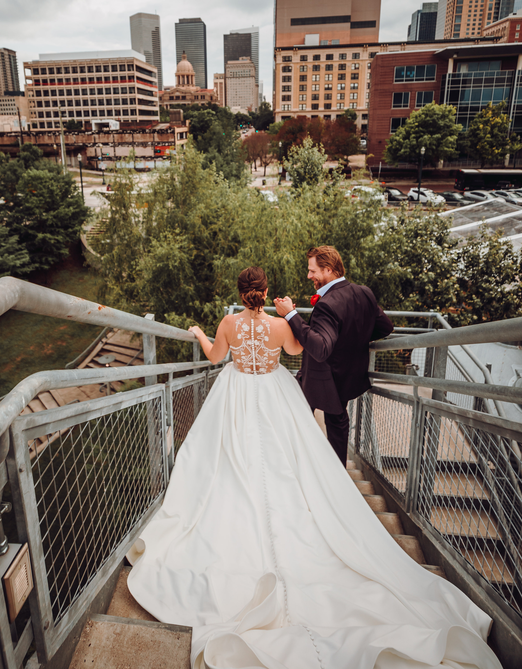 A groom holds his bride's hand as they walk down the stairs with Houston skyscrapers in the background.