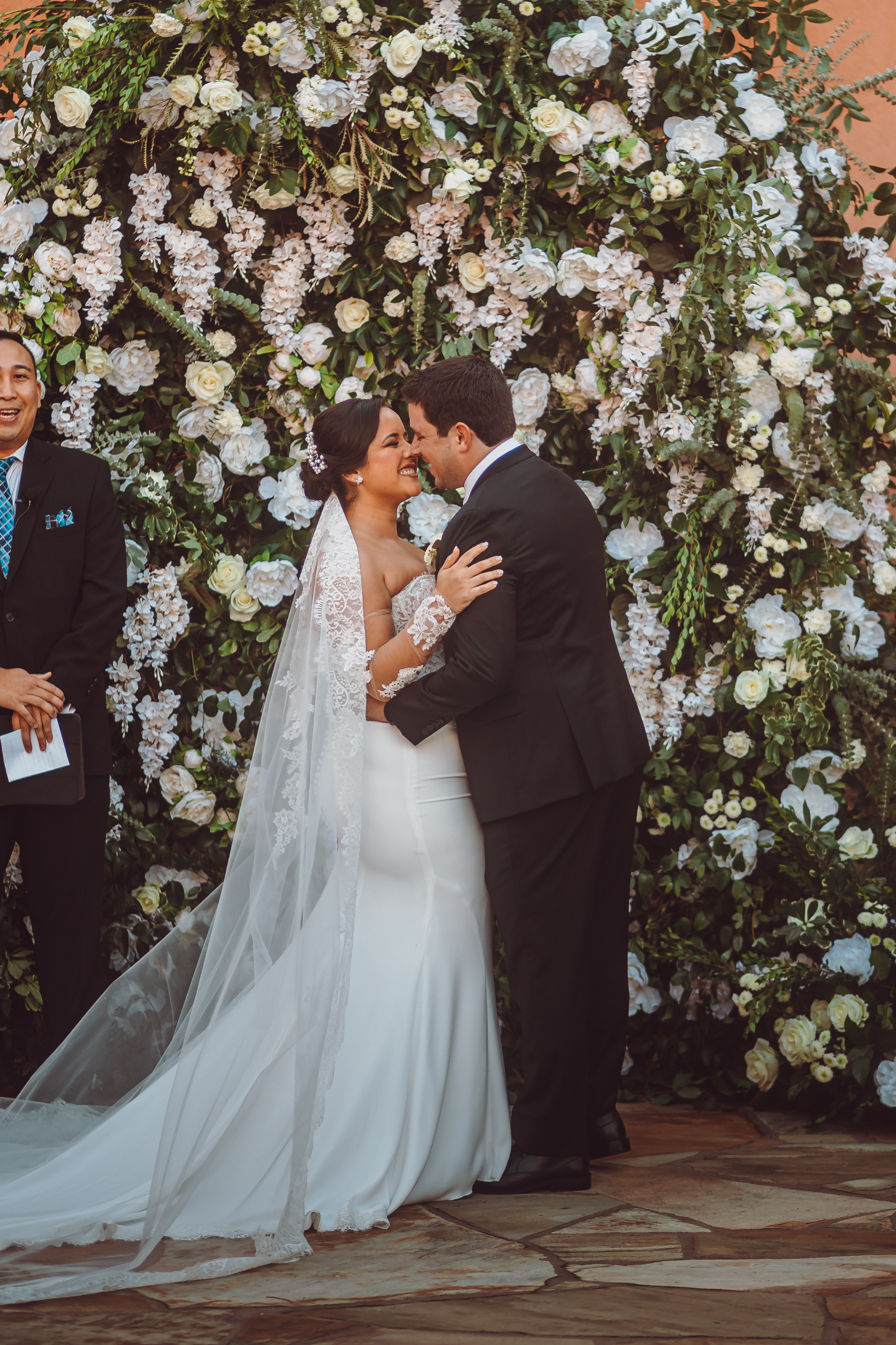 Groom holds a smiling bride at their outdoor wedding ceremony, aside a greenery and floral installation, at Agave Estates. 