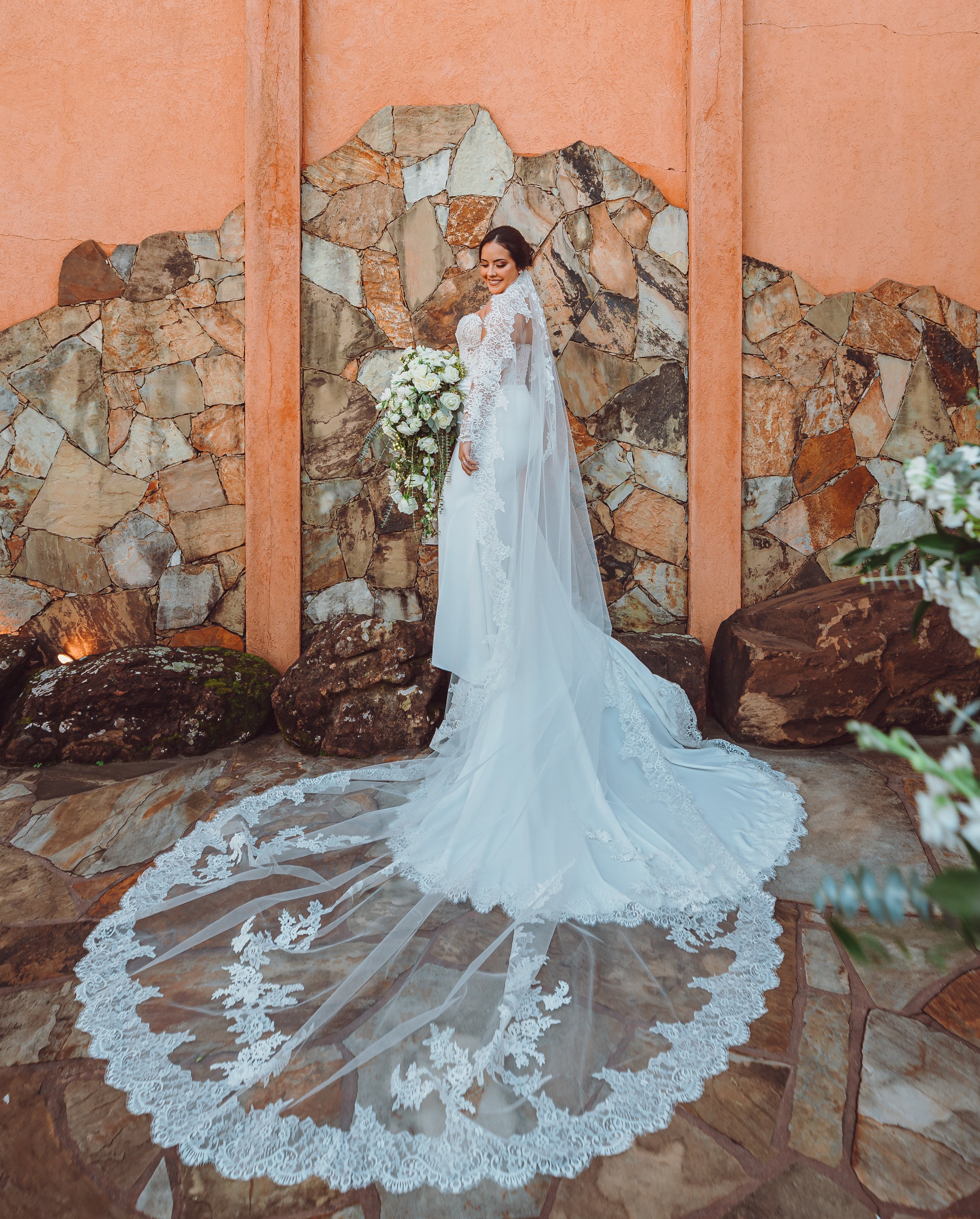 A bride stands against a terra cotta stone wall in her wedding dress. Her veil is spread around her.