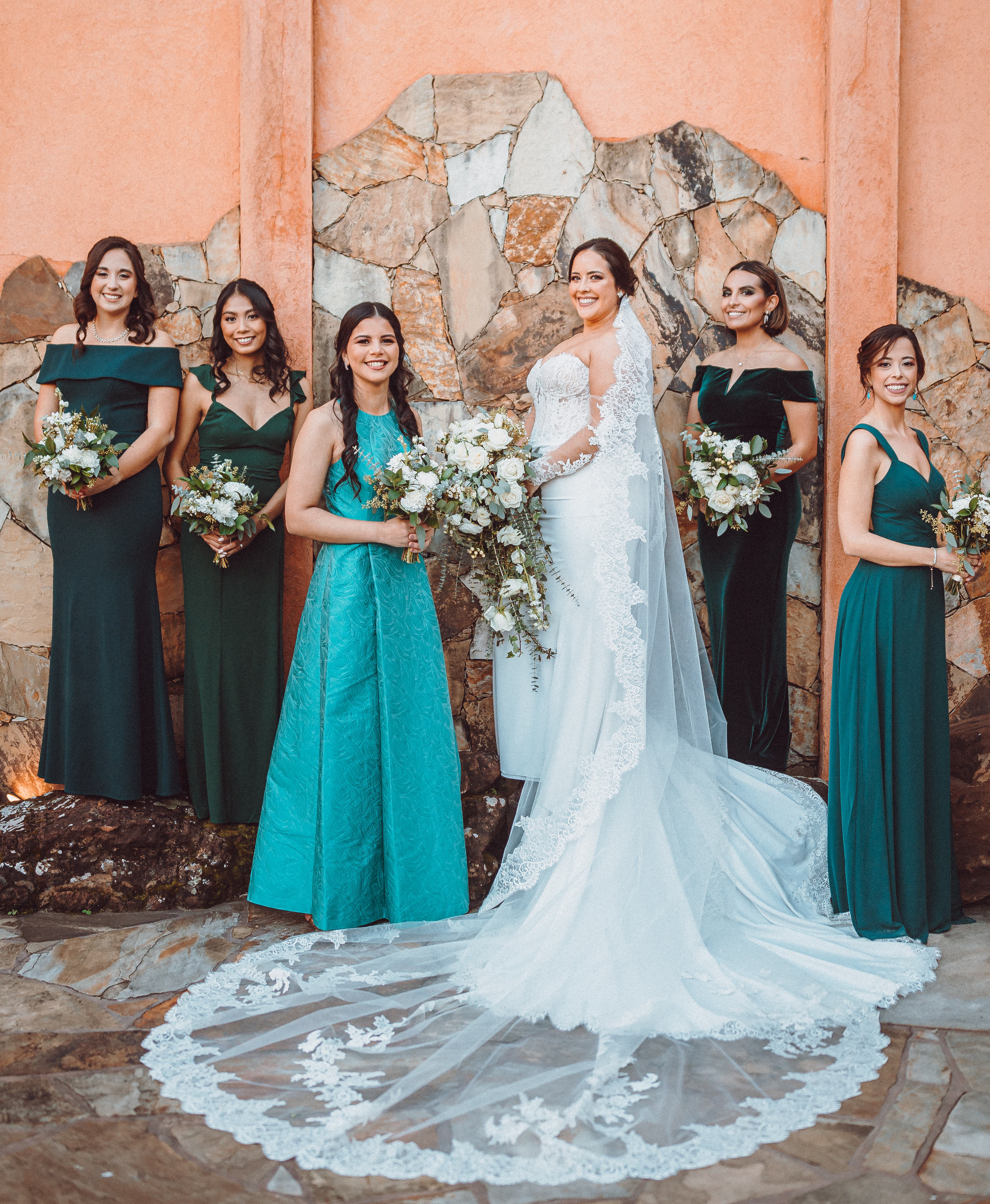 A bride stands against a terra cotta wall with her bridesmaids surrounding her in turquoise and green dresses.
