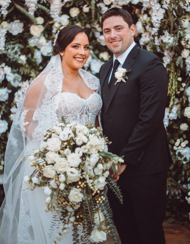 A smiling bride and groom stand together aside a greenery and white floral installation after their wedding ceremony at Agave Estates.