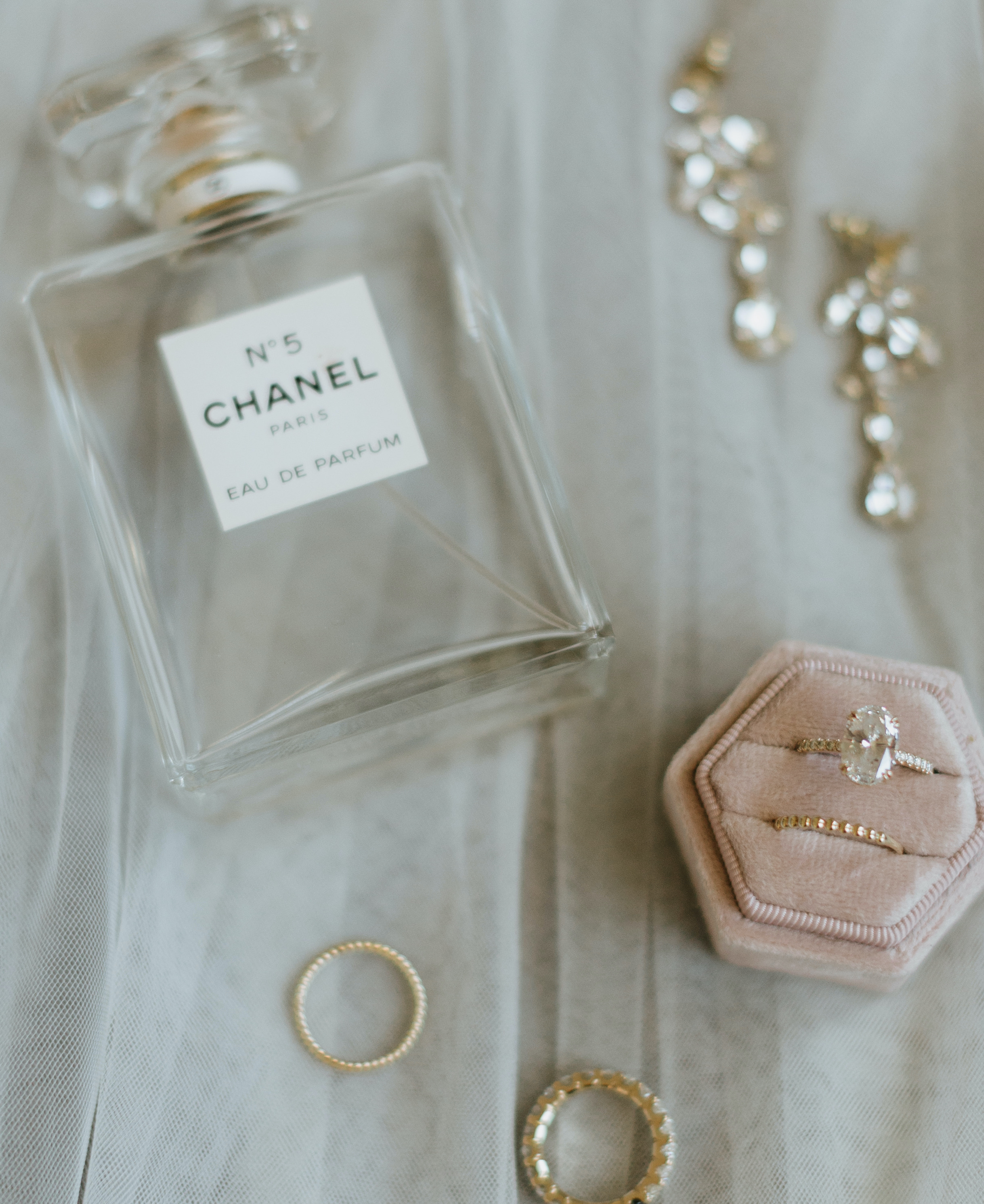 A flatlay of a Chanel perfume bottle and the bride's oval diamond engagement ring in a mauve pink ring box with her wedding band.