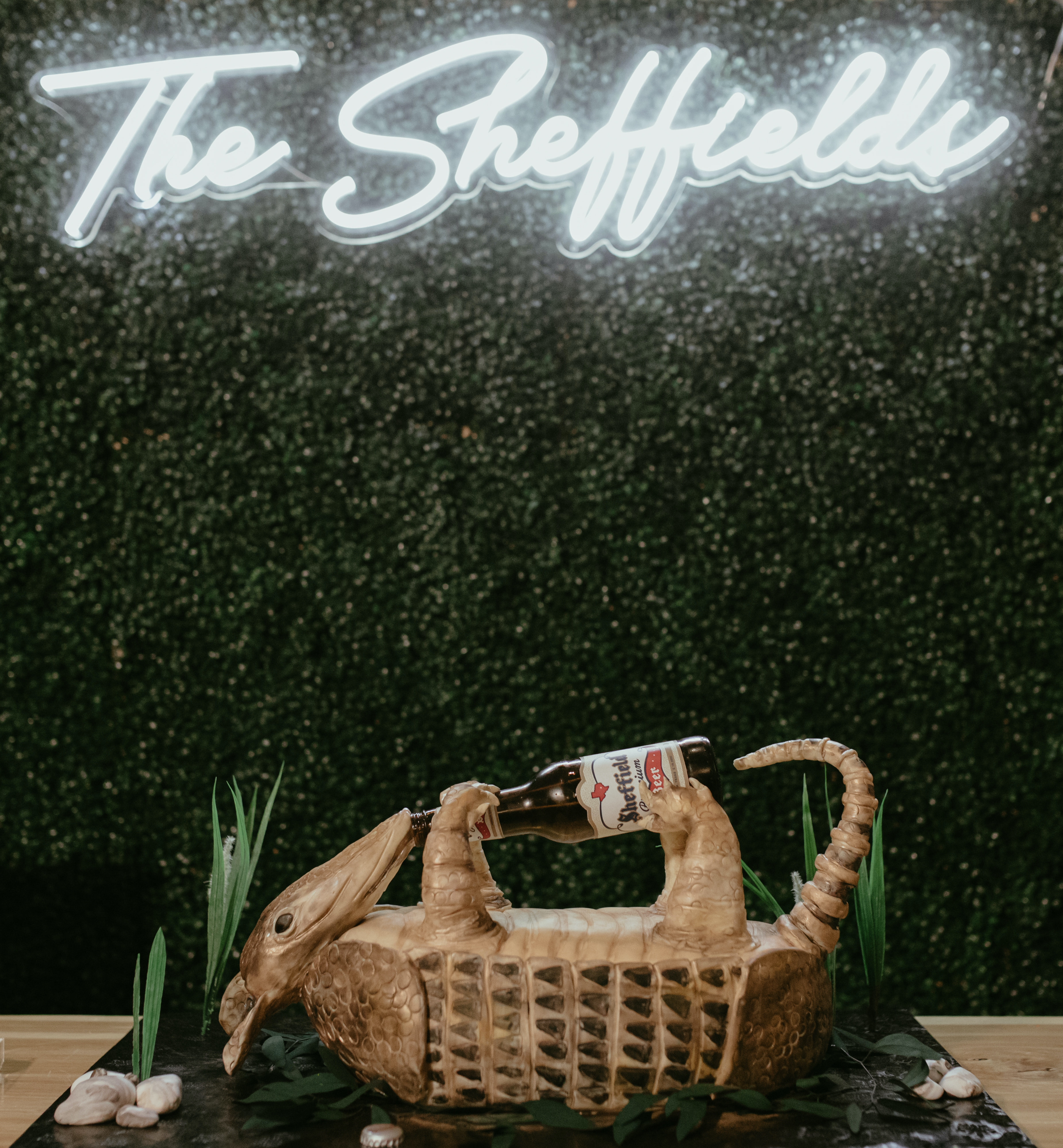 A neon sign for a wedding is hanging on a hedge wall. The groom's cake in the shape of an armadillo is sitting in front of it. Peach-Toned Countryside wedding.
