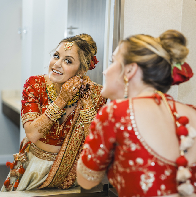 Bride in red and gold wedding saree looks at her reflection while putting on earrings before her hindu wedding ceremony at Marriott Cityplace at Springwoods Village. 