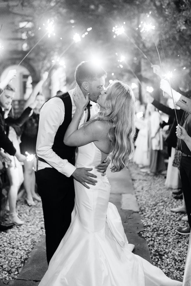 A black and white photo of the bride and groom kissing under sparklers as they exit the wedding reception. 