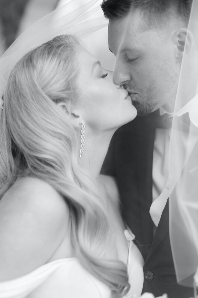A black and white photo of a bride and groom sharing a kiss under the bride's vale. 
