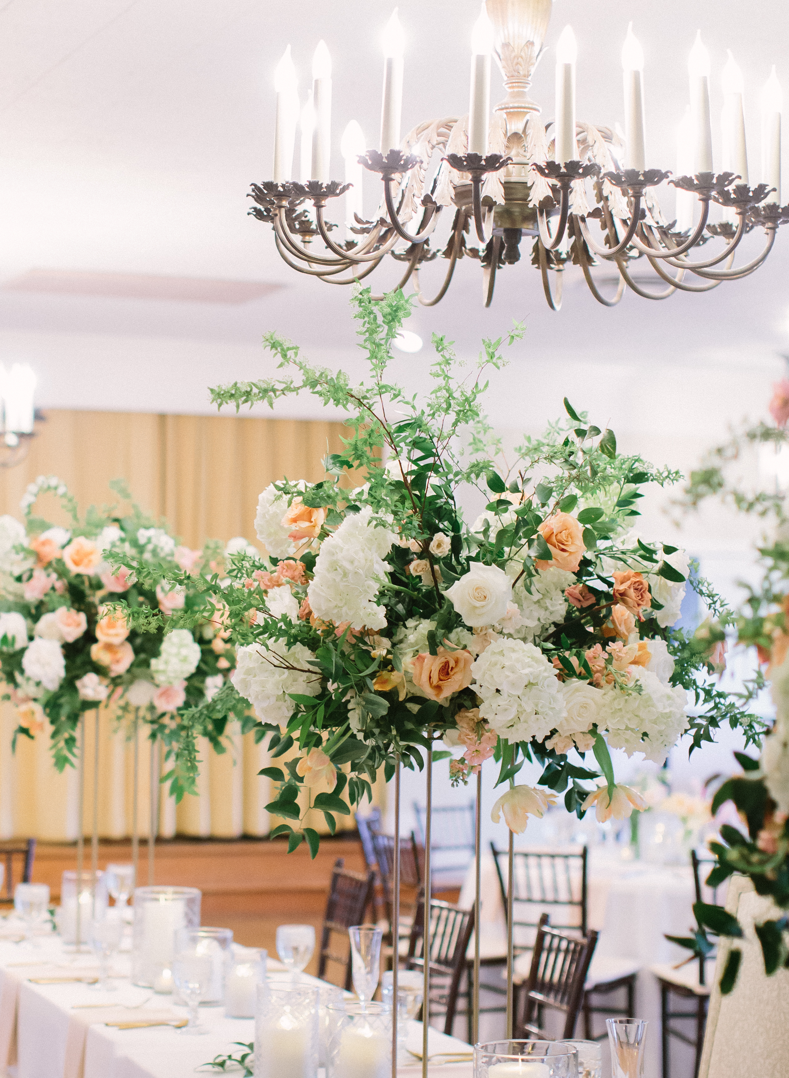 A wedding reception room is decorated with greenery and tall floral centerpieces with cream, peach and pink flowers for an early spring romance wedding.