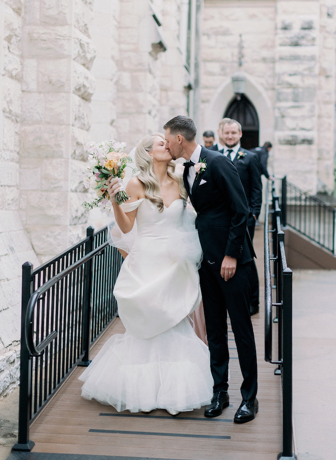 A bride while holding her bouquet kisses the groom as they exit their wedding ceremony outside of the church. 