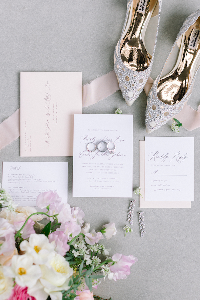 A wedding invitation suite, pink envelope with cursive font, blush colored ribbon, a brides floral bouquet and pair of Badgley Mischka pumps.