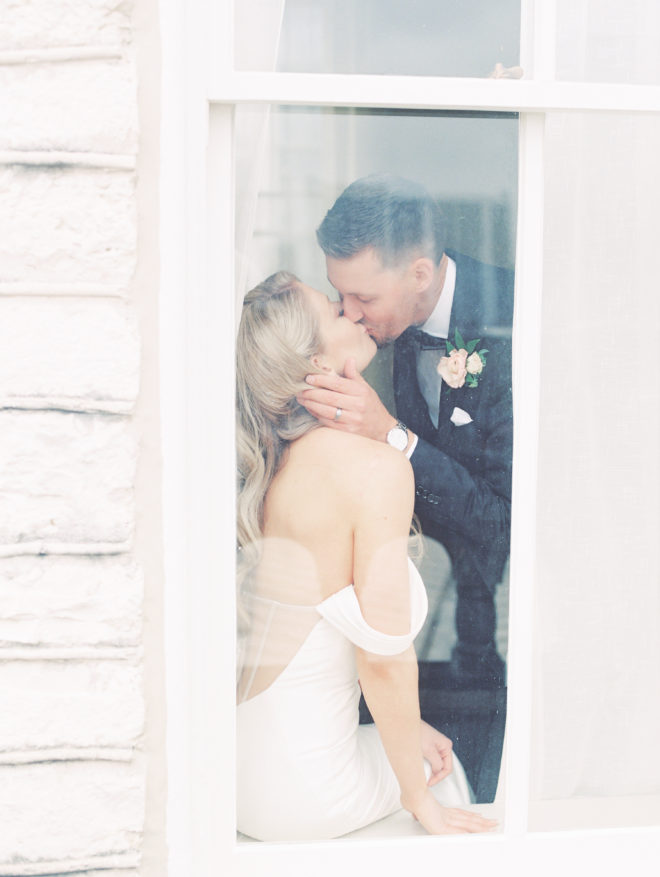 A bride sits on a windowsill while a groom leans down to kiss her on their wedding day in Austin, TX.