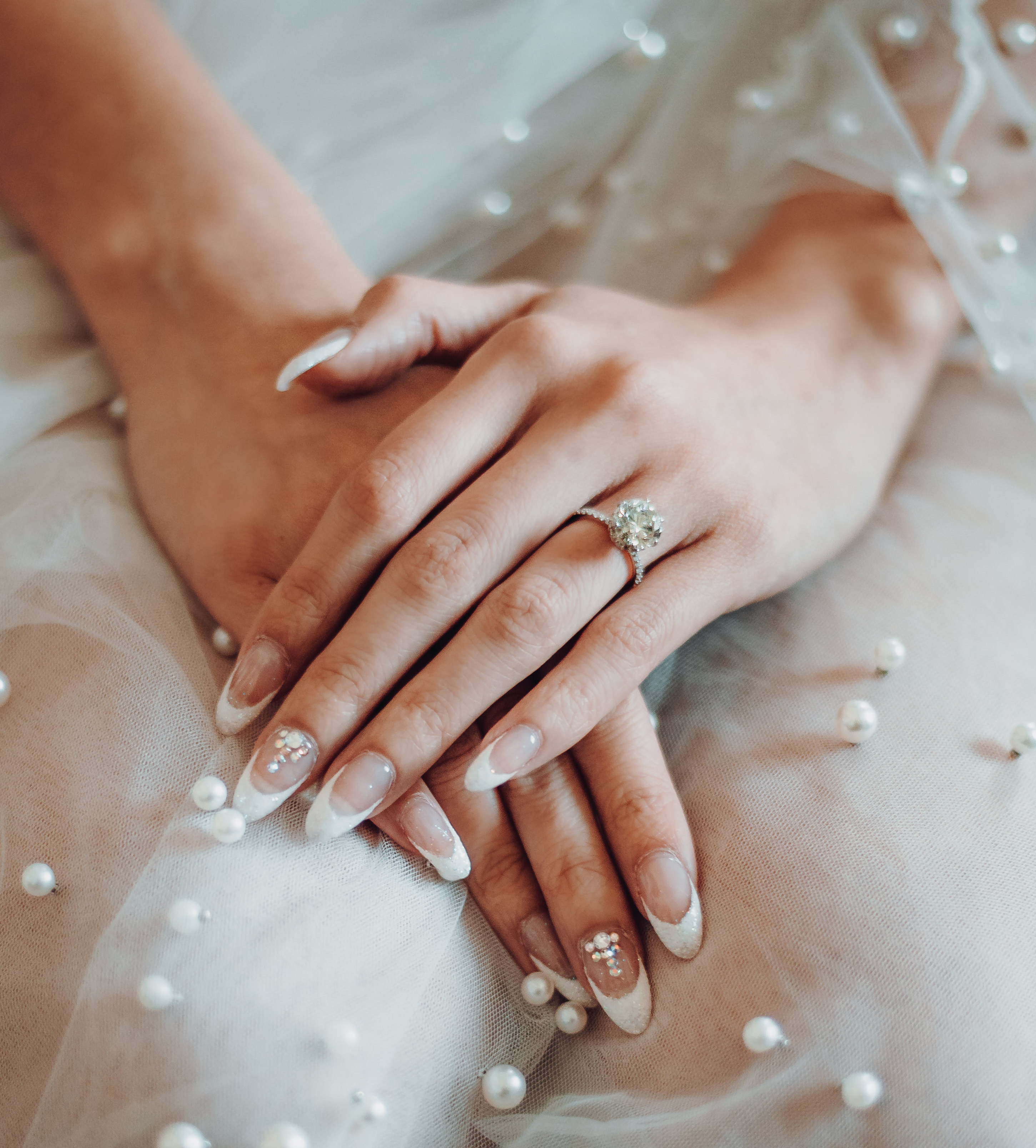 A bride's freshly manicured hands and her diamond engagement ring are placed on her lap. 