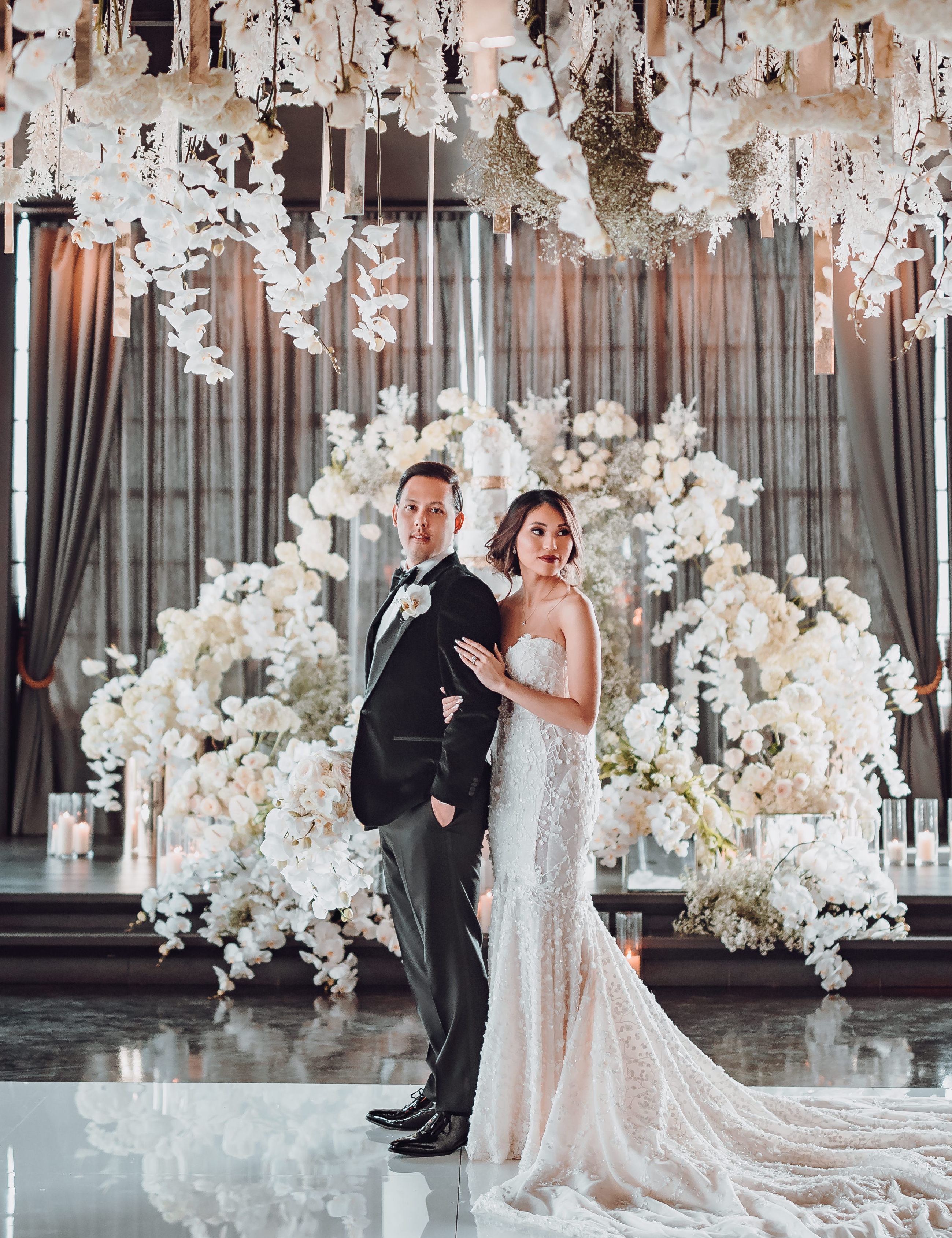 A groom in a black tuxedo stands with a bride in a Galia Lahav wedding gown in the Astorian's ballroom, decorated with white floral installations and lit candles.
