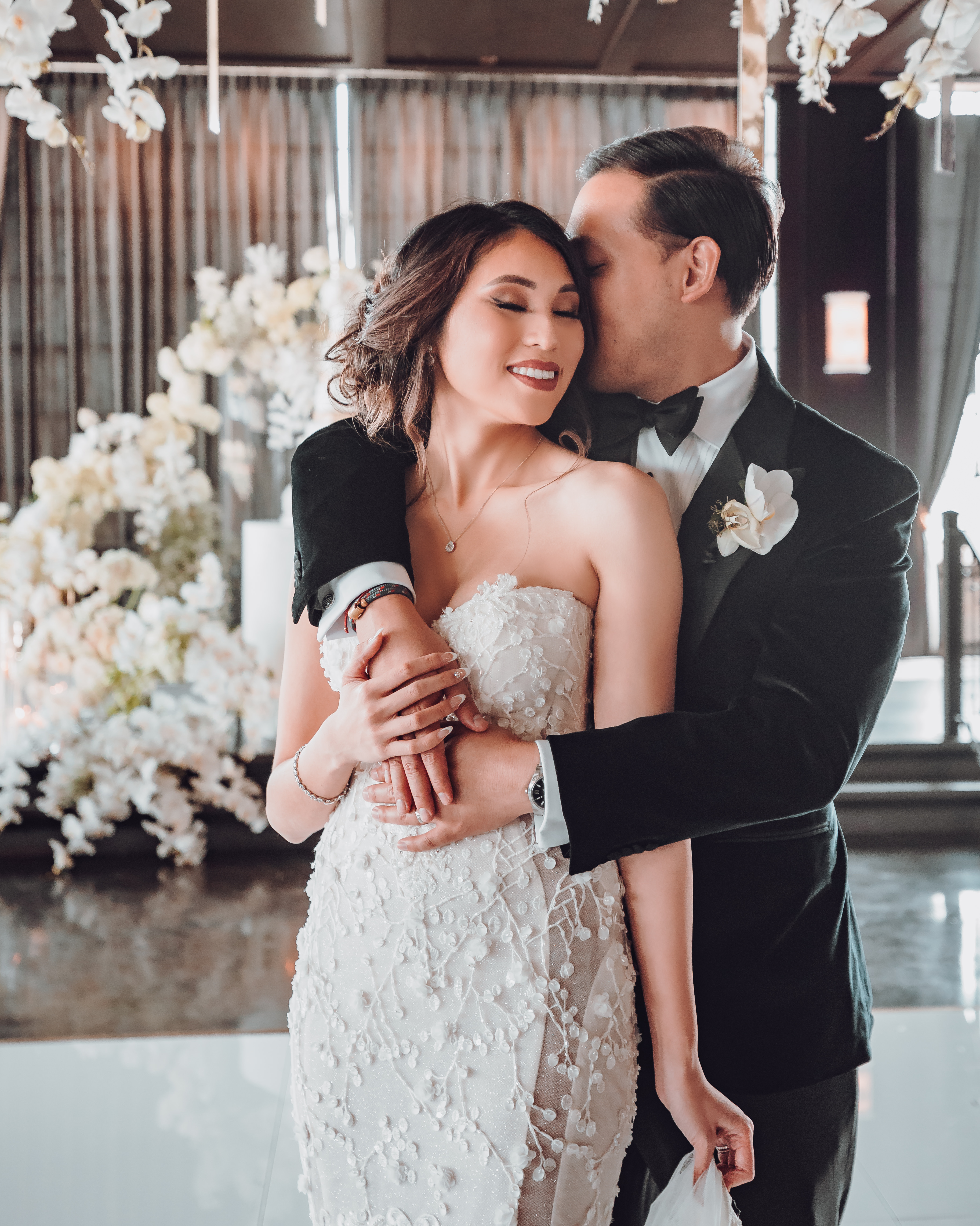 A groom hugs his bride from behind as she closes her eyes and smiles. They are standing in the reception room of their wedding venue The Astorian in Houston, TX.