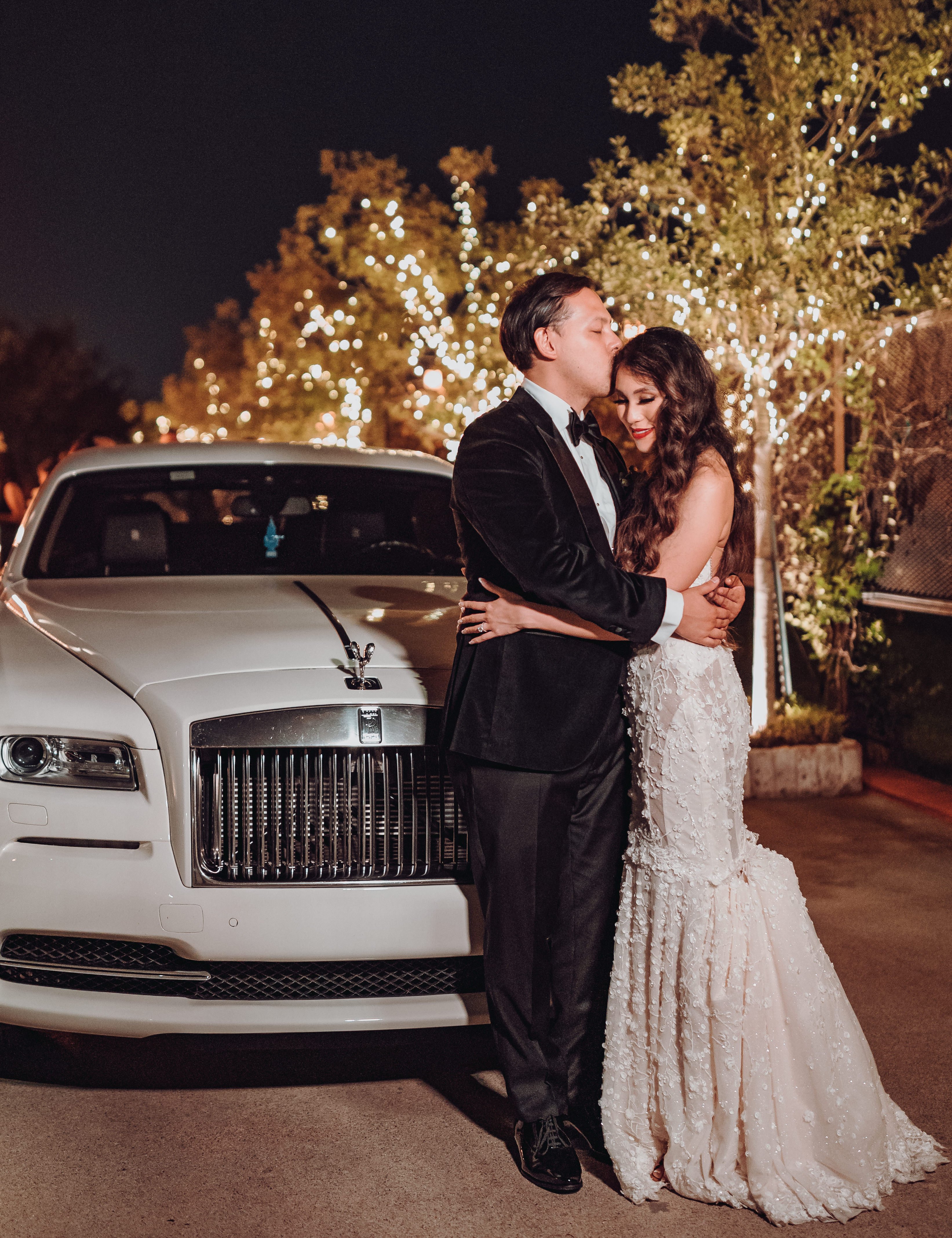 A groom kisses his bride on the cheak as they stand outside their getaway car which is a Rolls Royce.