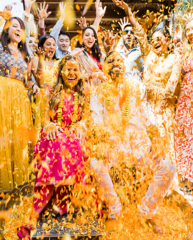 Bride and groom smile as guests throw marigold flowers at their Hindu celebration.