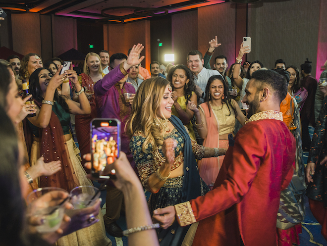 Blonde bride in teal and red indian bridal attire dances with a groom in a red kurta in a ballroom at Marriott Cityplace at Springwoods Village. 
