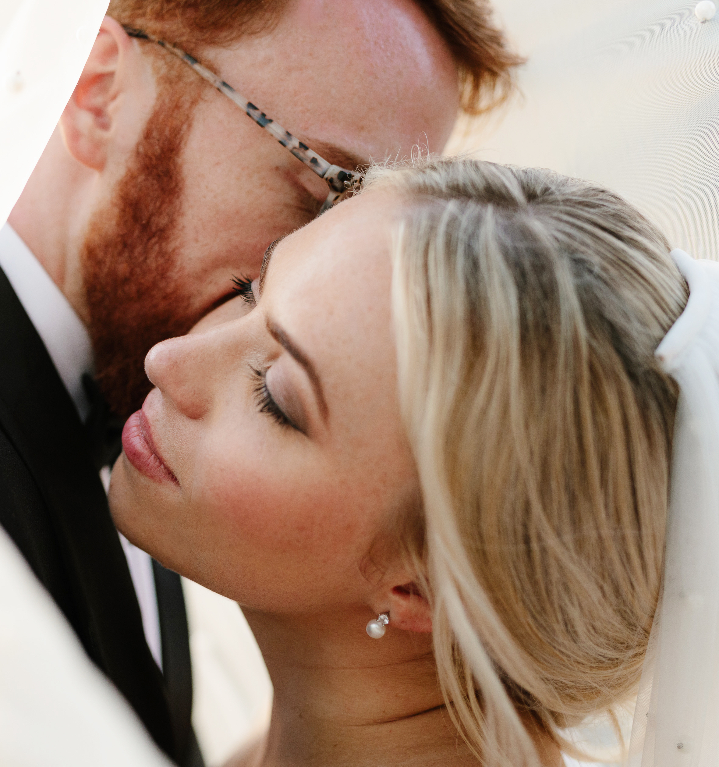 A close up photo of a groom kissing his bride's cheek as her veil wraps around them in the wind.