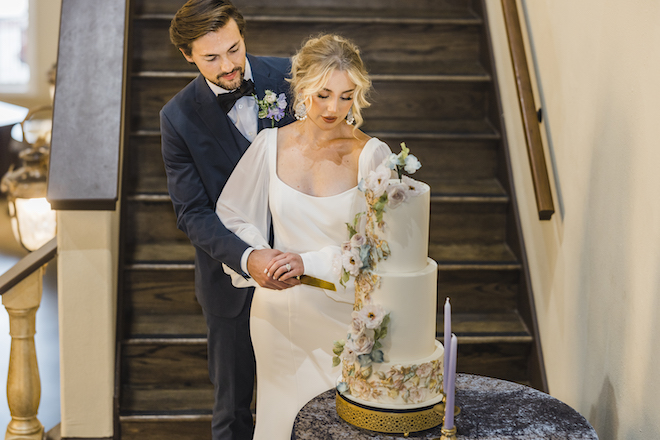 Groom in blue suit and bride in square neck wedding gown cut a 3-tier white wedding cake with lilac, pink and green flowers created by Chocolate Llama.