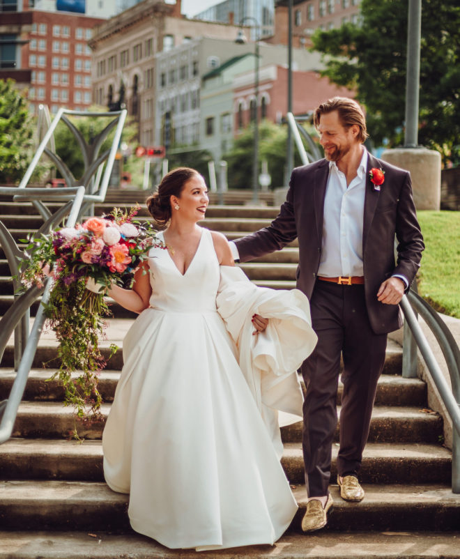 Bride holding a lush bouquet of flowers walks down the stairs with a groom in gold driving shoes outside of the Sunset Coffee Building in Houston, Texas.