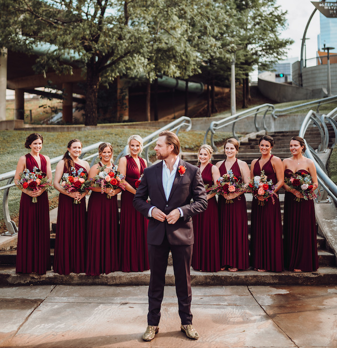 The groom stands in front of the bridesmaids who are wearing burgundy bridesmaid dresses and holding their bouquets. 