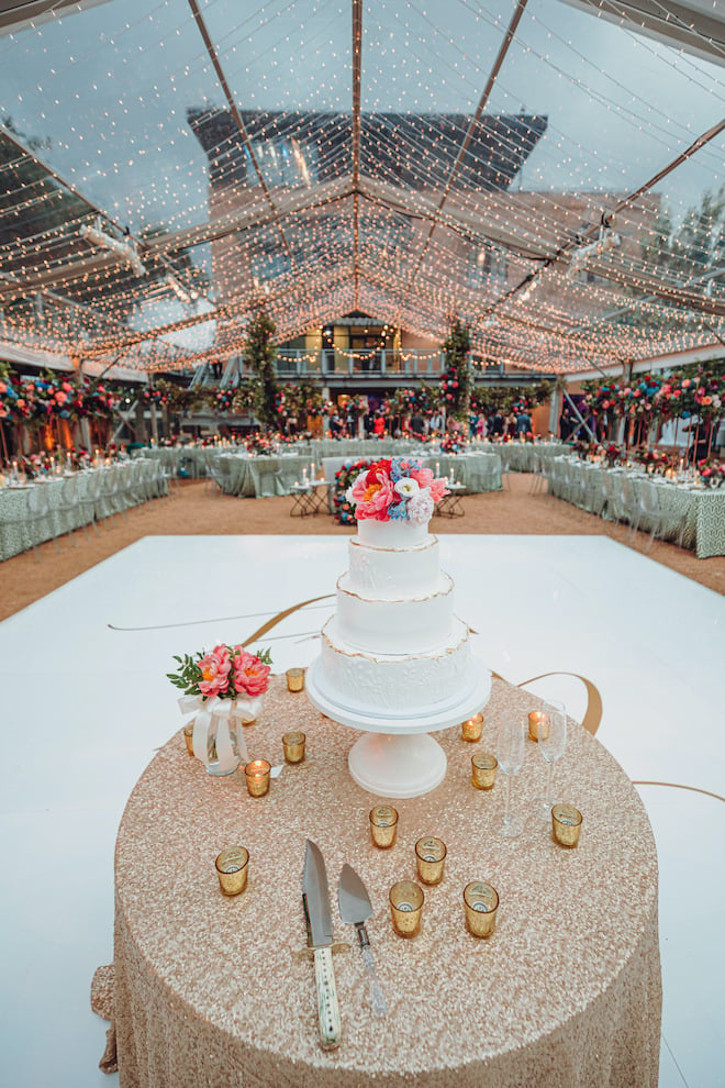 A 4-tier wedding cake sits on a white cake stand on the table outside under a canopy of string lights. 