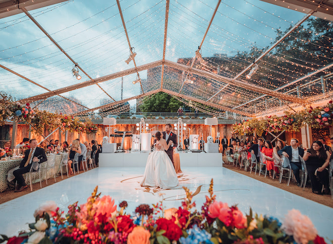 A bride and groom dance outside on the dance floor under a canopy of lights as guests watch. 