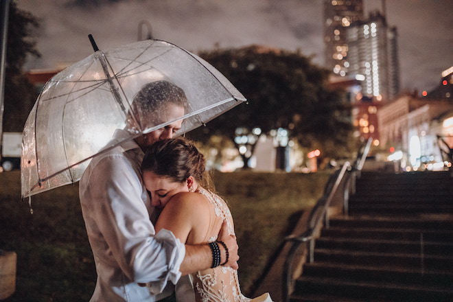 A bride and groom hug under an umbrella in the rain with the Houston skyline in the background. 