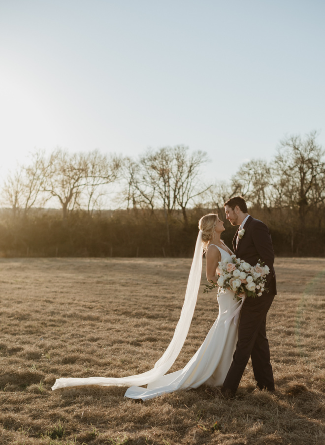 The bride and groom stand in an open field outside their Texas wedding venue, Deep In The Heart Farms. 