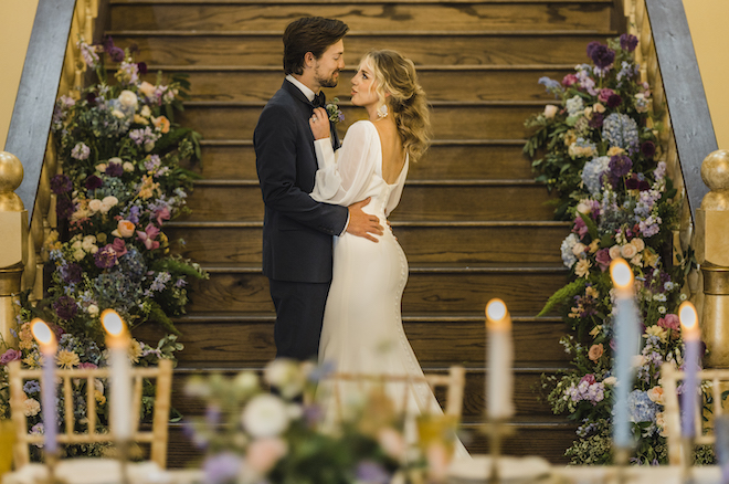 The bride and groom smile at the bottom of the stairs looking at each other surrounded by purple, blue and peach flowers at their wedding reception. 