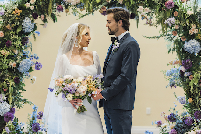A bride and groom stand holding hands under a floral arch of blue, peach, pink and purple flowers. 