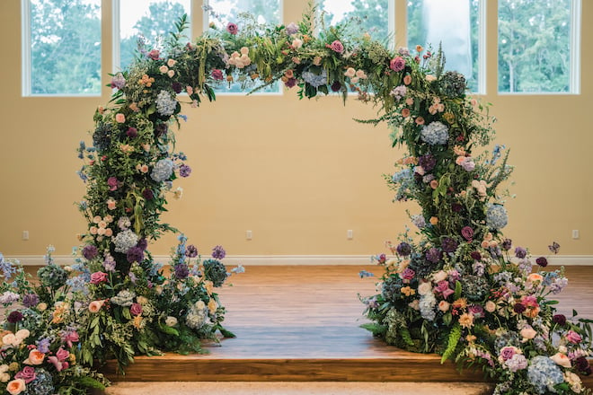 An arch of purple, blue, peach and cream flowers for the wedding ceremony at the venue, The Hundred Oaks. 