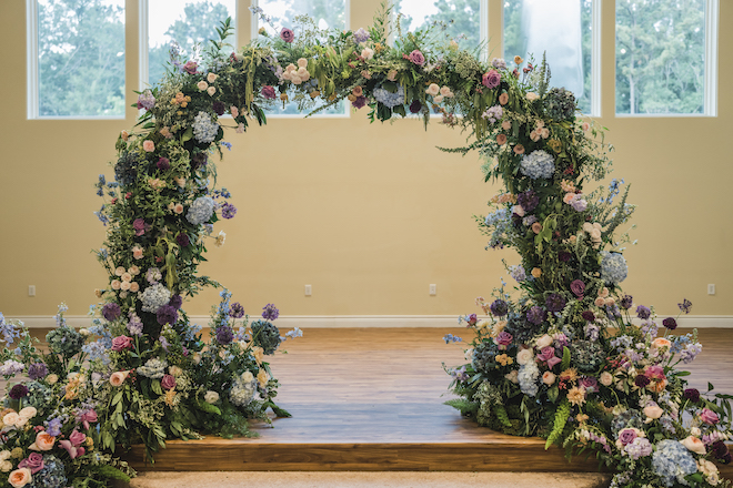 An arch of purple, blue, peach and cream flowers for the wedding ceremony at the venue, The Hundred Oaks. 