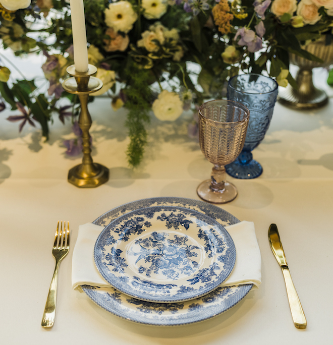 White and blue floral, fine china are placed aa the table next to gold silverware and pink and blue glasses. 