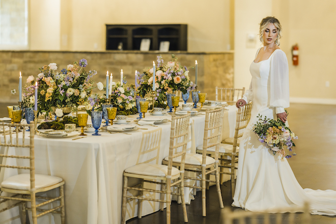 A bride stands by a table decorated with candles and blue, pink, peach and purple flowers at the wedding venue, The Hundred Oaks. 