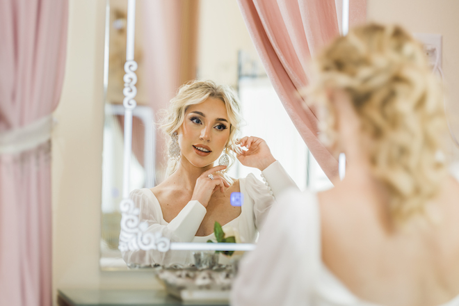 A bride wearing a wedding dress looks in the mirror as she puts on silver diamond earrings. 
