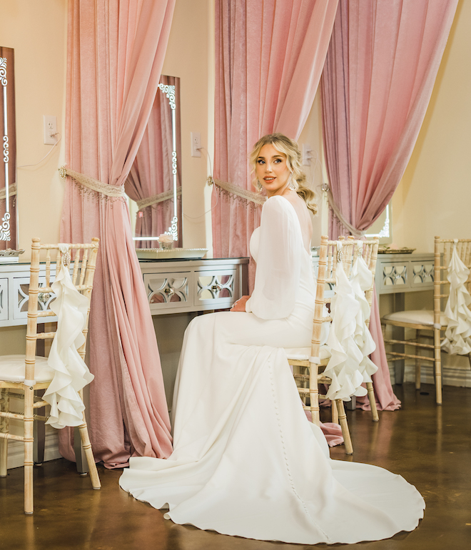 A bride sitting down in a chair wearing her wedding gown looks over her shoulder with her hair and makeup done. 