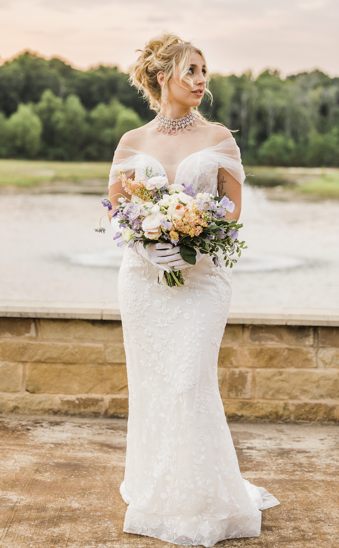 A bride wears an off-the-shoulder, white lace wedding dress from Parvani Vida while holding a bouquet of garden-inspired florals by Bloom & Grove. 