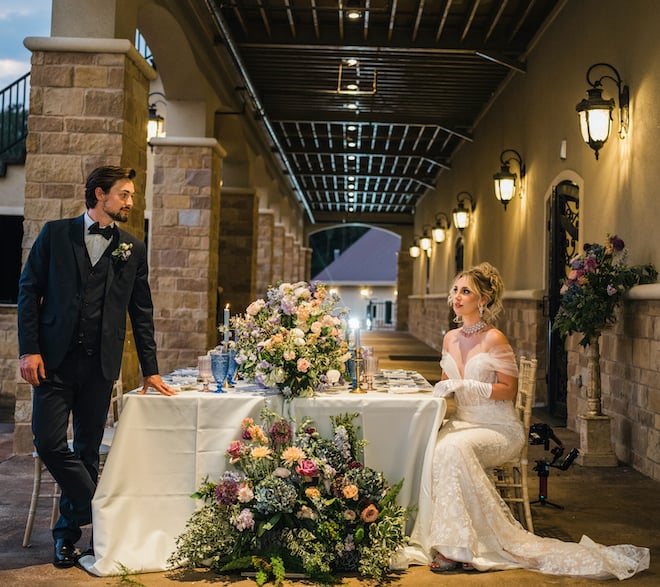Groom leans on a family-style table decorated with flowers, warm candlelight and tableware, while a bride sits at a chivari chair at a wedding editorial at The Hundred Oaks wedding venue in Houston, TX.