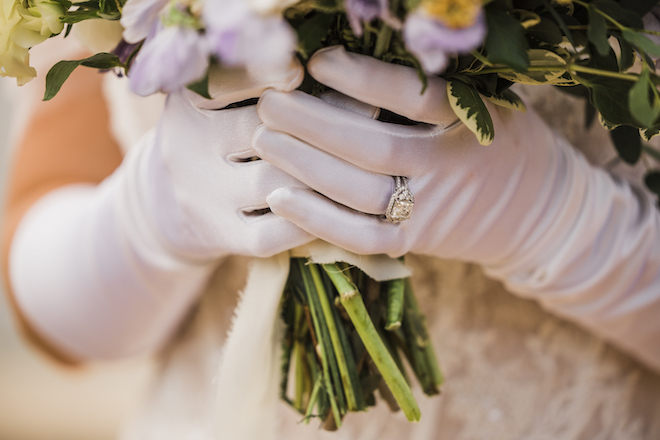 A bride wears white, silk gloves with her wedding ring on as she holds wedding bouquet. 
