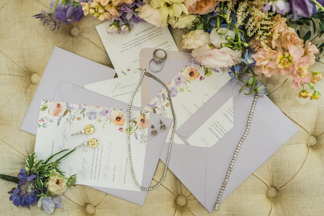 Purple, pink and yellow flowers surround a wedding invitation suite and fine bridal jewelry at a wedding styled shoot at venue, The Hundred Oaks.