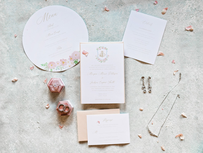 Fine jewelry lay next to blush pink wedding stationery and invitations and blush pink rose petals. 