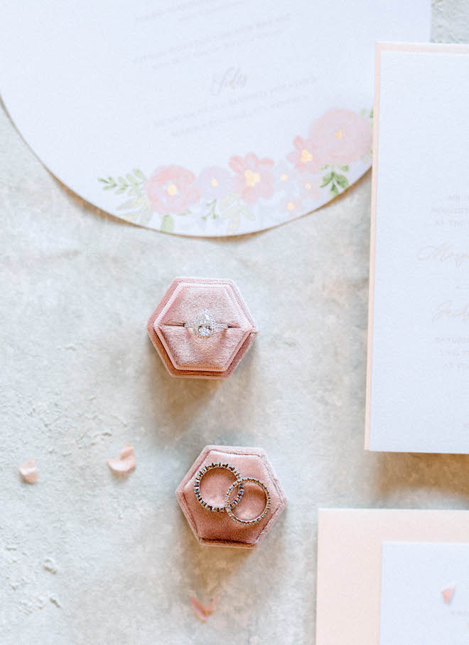 A wedding ring and wedding bands lay on pink and velvet ring cases next to blush pink stationery. 