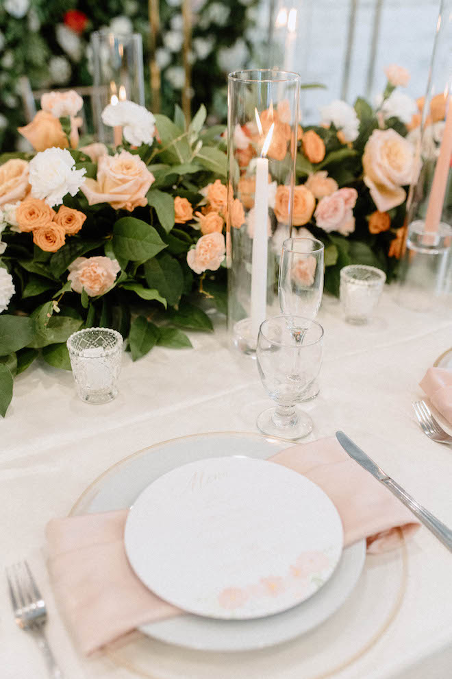 The table is decorated with fine china, blush pink napkins, candles, white linens and peach, white and pink flowers. 