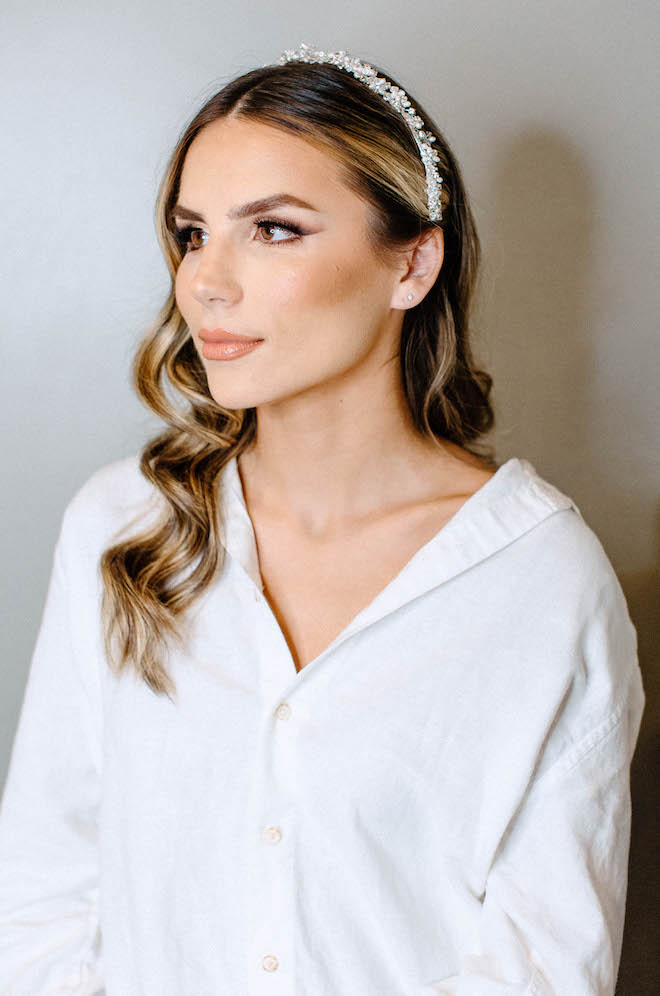 A bride sits wearing a white button up and a sparkling head band with her hair curled and makeup done. 