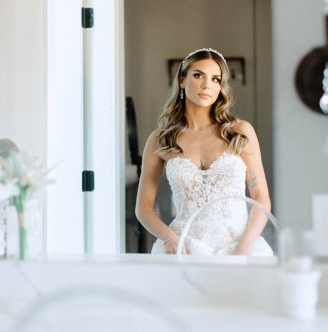 The bride looks in the mirror while wearing a lace, strapless wedding gown and diamond jewelry. 