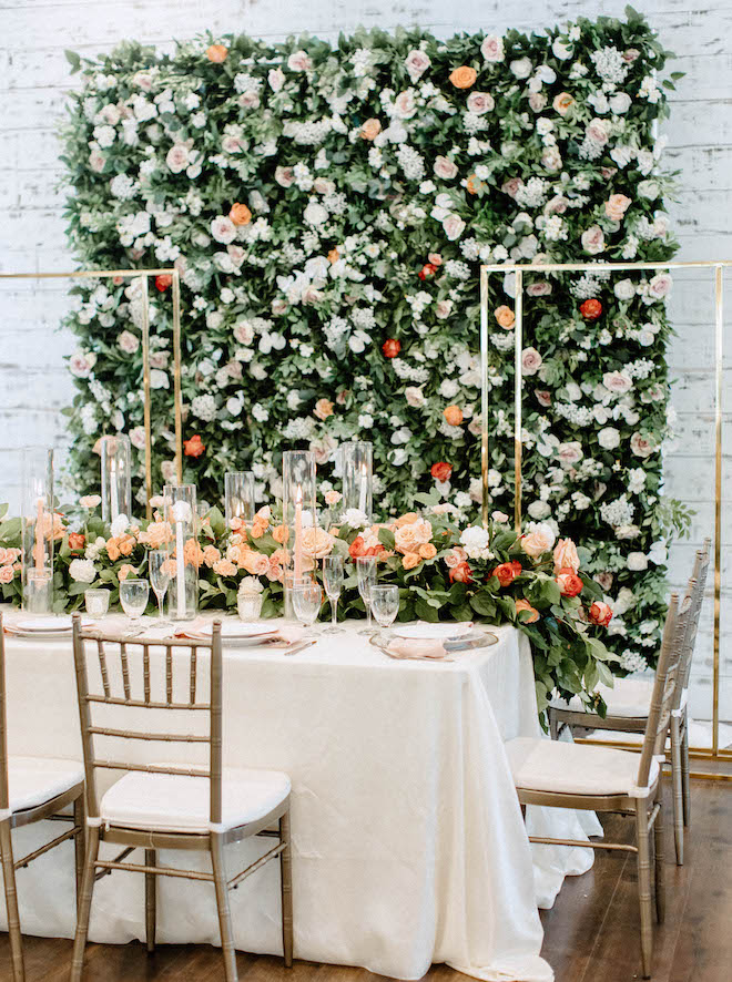 A table with orange, peach , pink and red flowers sits in front of a flower wall decorated with white, orange and red flowers at the wedding venue, Mara Villa. 